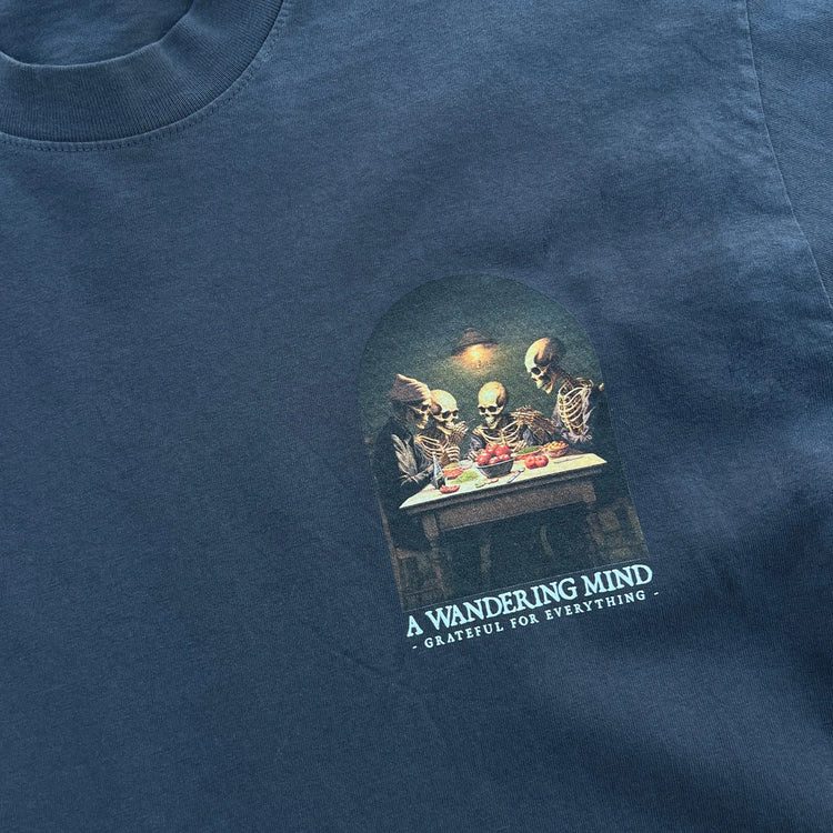 A Wandering Mind Grateful for Everything t-shirt in slate front graphic detail showing skeletons at table, food, interior scene