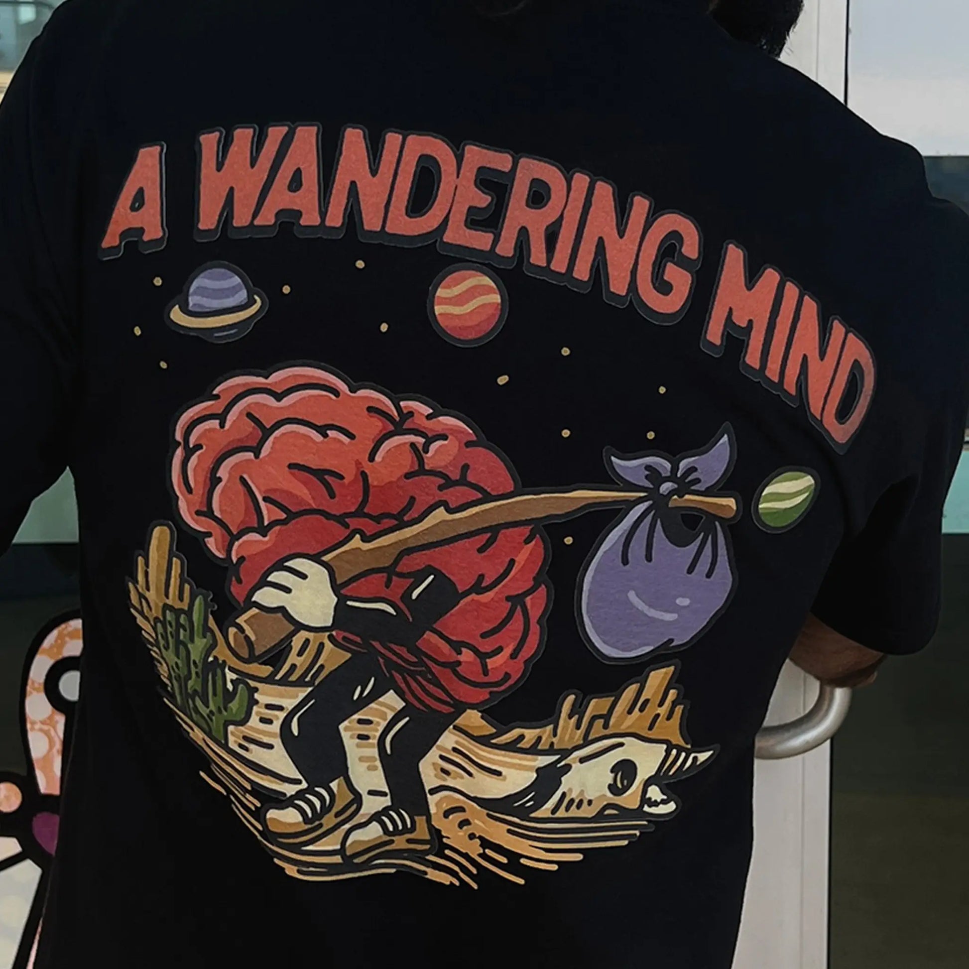 A Wandering Mind Arid Thoughts t-shirt in black back graphic featuring wandering brain character, desert, skull