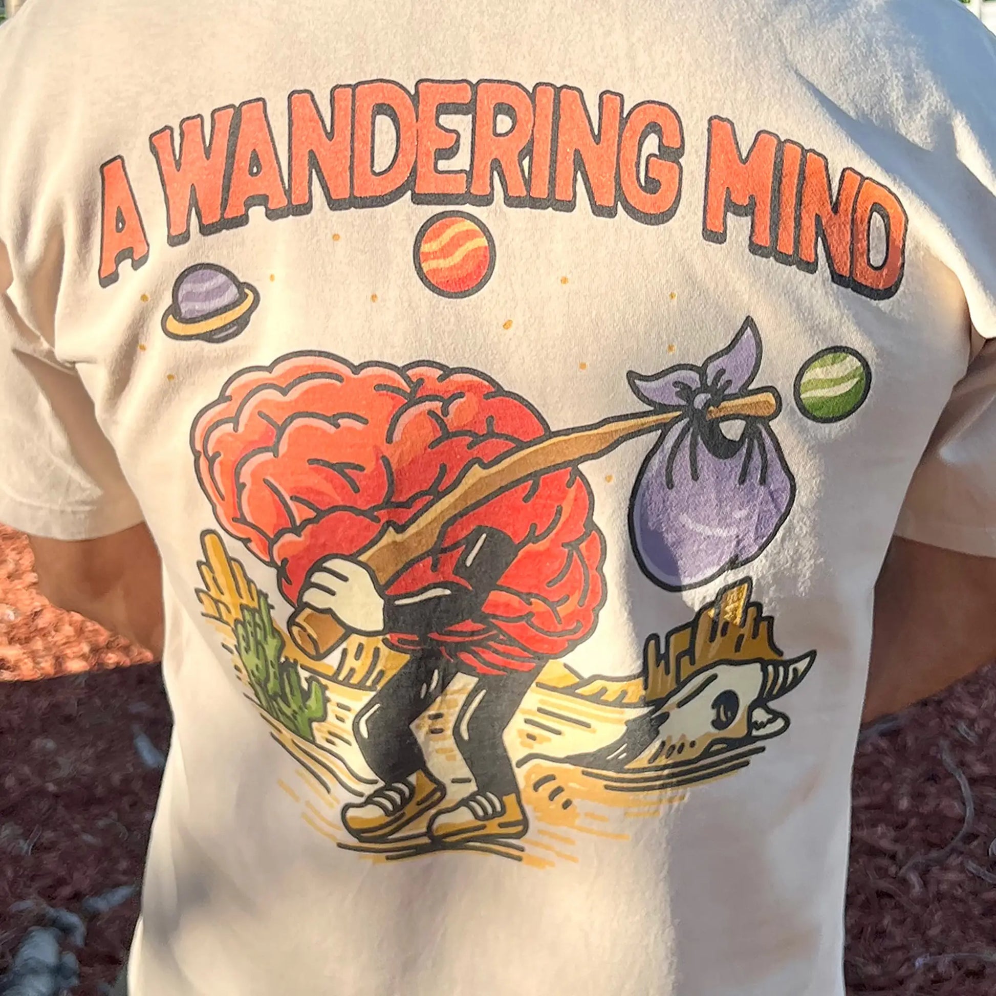A Wandering Mind Arid Thoughts t-shirt in sand back graphic featuring wandering brain character, desert, skull